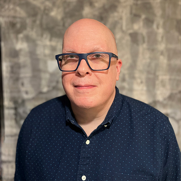 Headshot of Jeff, a bald man wearing blue glasses and a dark button-up shirt, smiling slightly against a neutral patterned gray background.