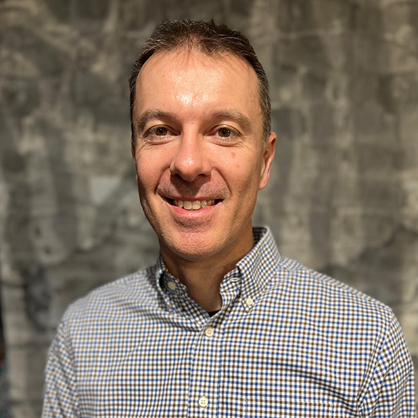Headshot of Michele, a man with short brown hair wearing a light checkered button-up shirt, smiling in front of a neutral patterned gray background.